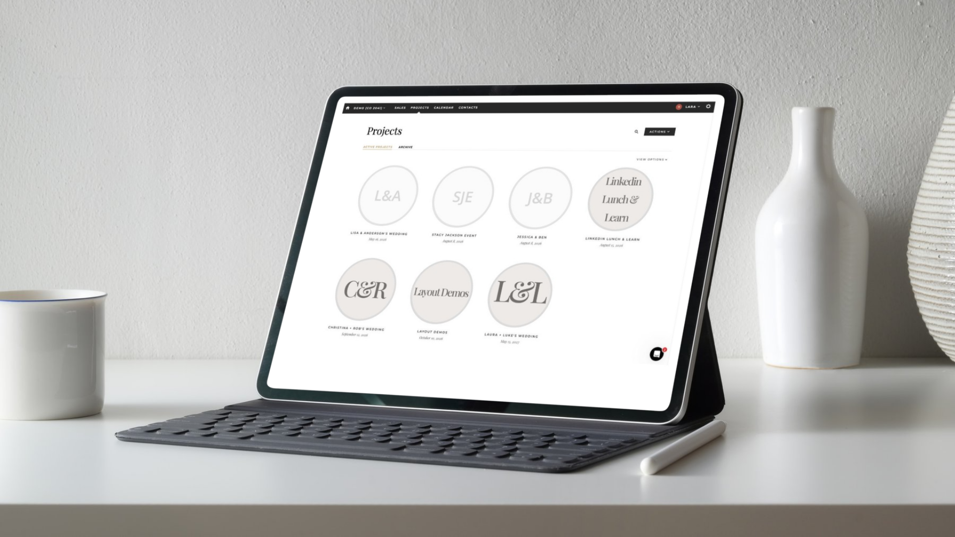 A professional tablet with a keyboard displays the Aisle Planner Pro wedding planning software and portfolio on a white desk, alongside a coffee mug and digital stylus for a modern event coordinator workspace.
