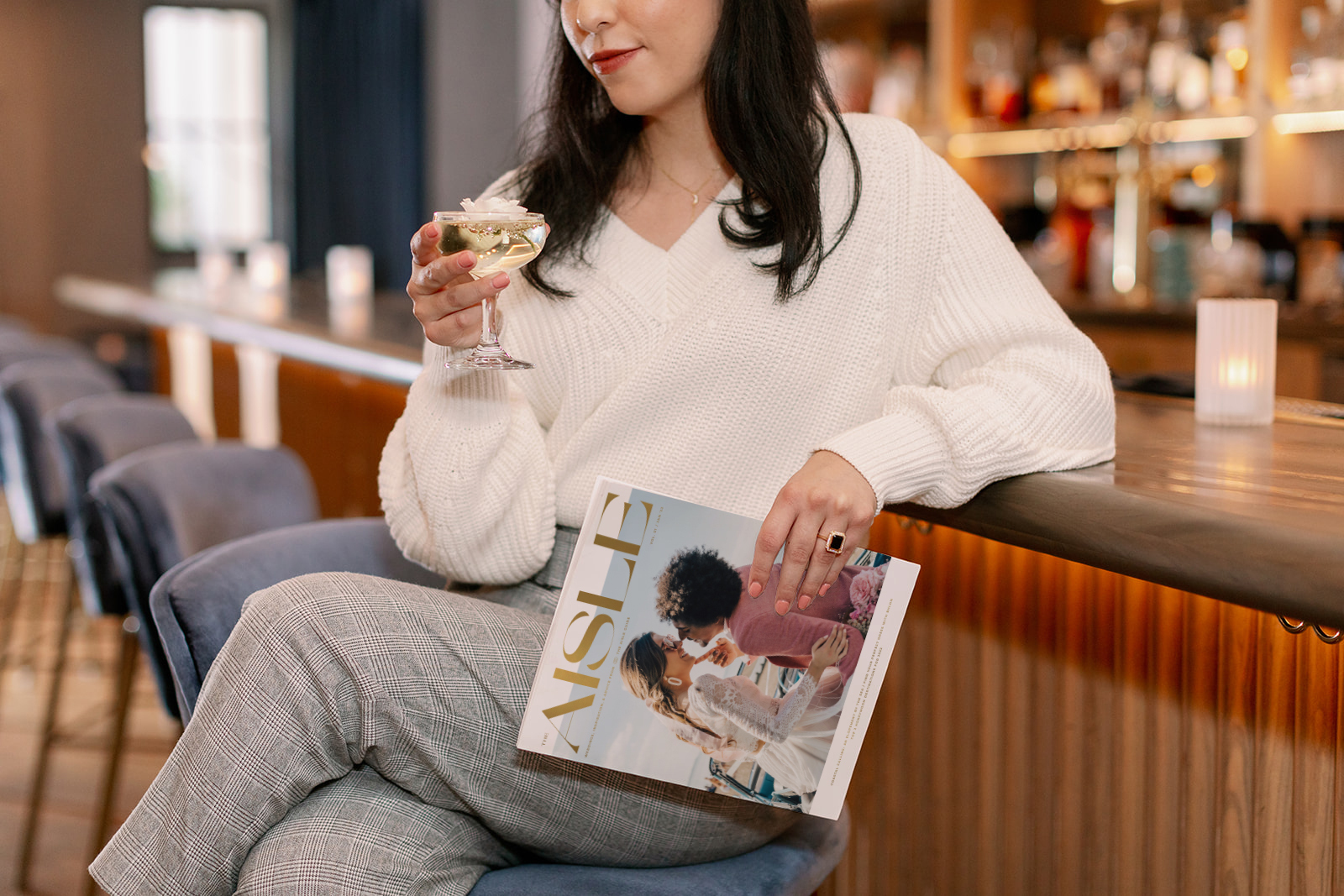 A woman at a bar with The Aisle Magazine and a drink in her hand 