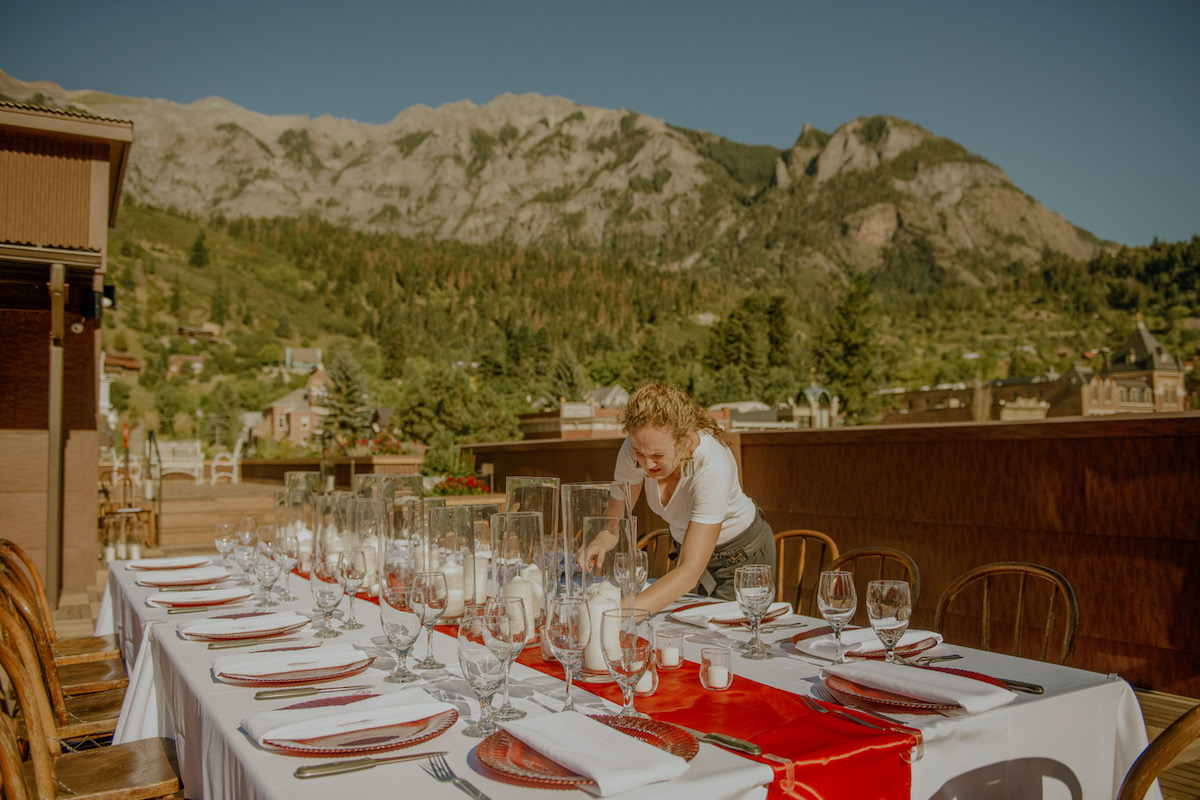 Krysta setting table with white tablecloth and bright red runner, green mountainous backdrop