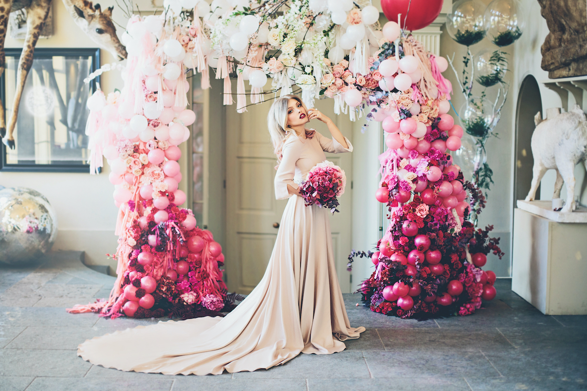 styled shoot with bride wearing a flowing gown and holding a pink bouquet under a pink, balloon filled archway