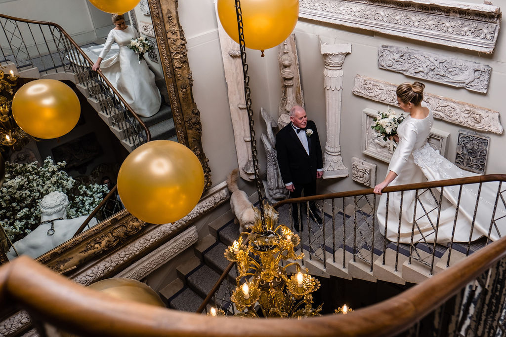Bride walking down ornate staircase to her father, holding a bouquet and surrounded by large gold balloons