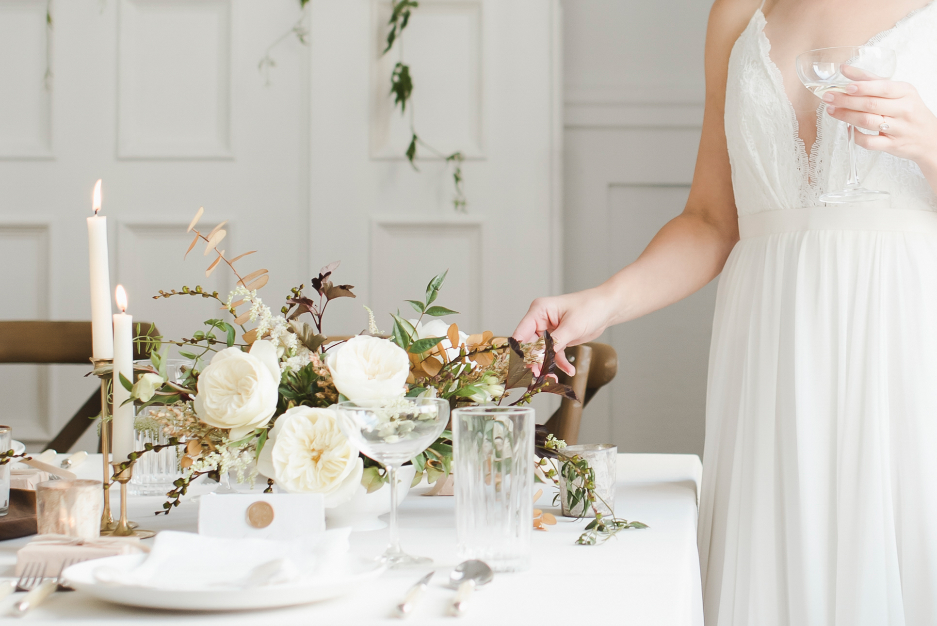 A person in a white dress setting a table with pink taper candles and a place setting with white dishes and glasses 