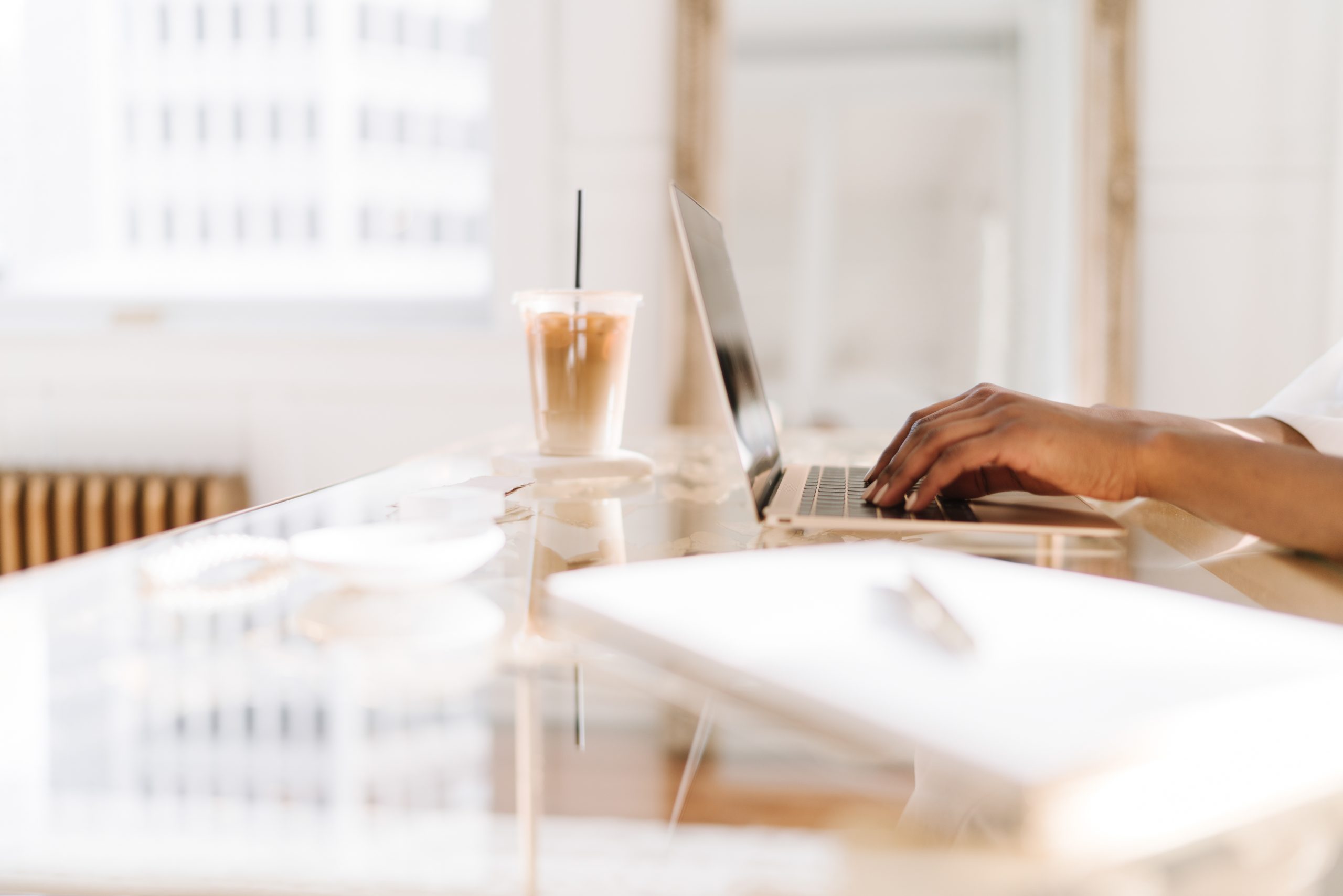 woman typing at computer with iced coffee and notebook on desk 