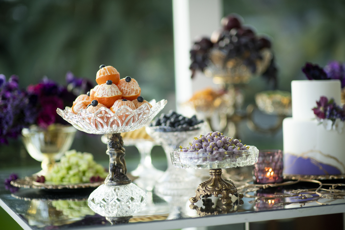 fruits and sweets laid out on table with lush green plant filled background