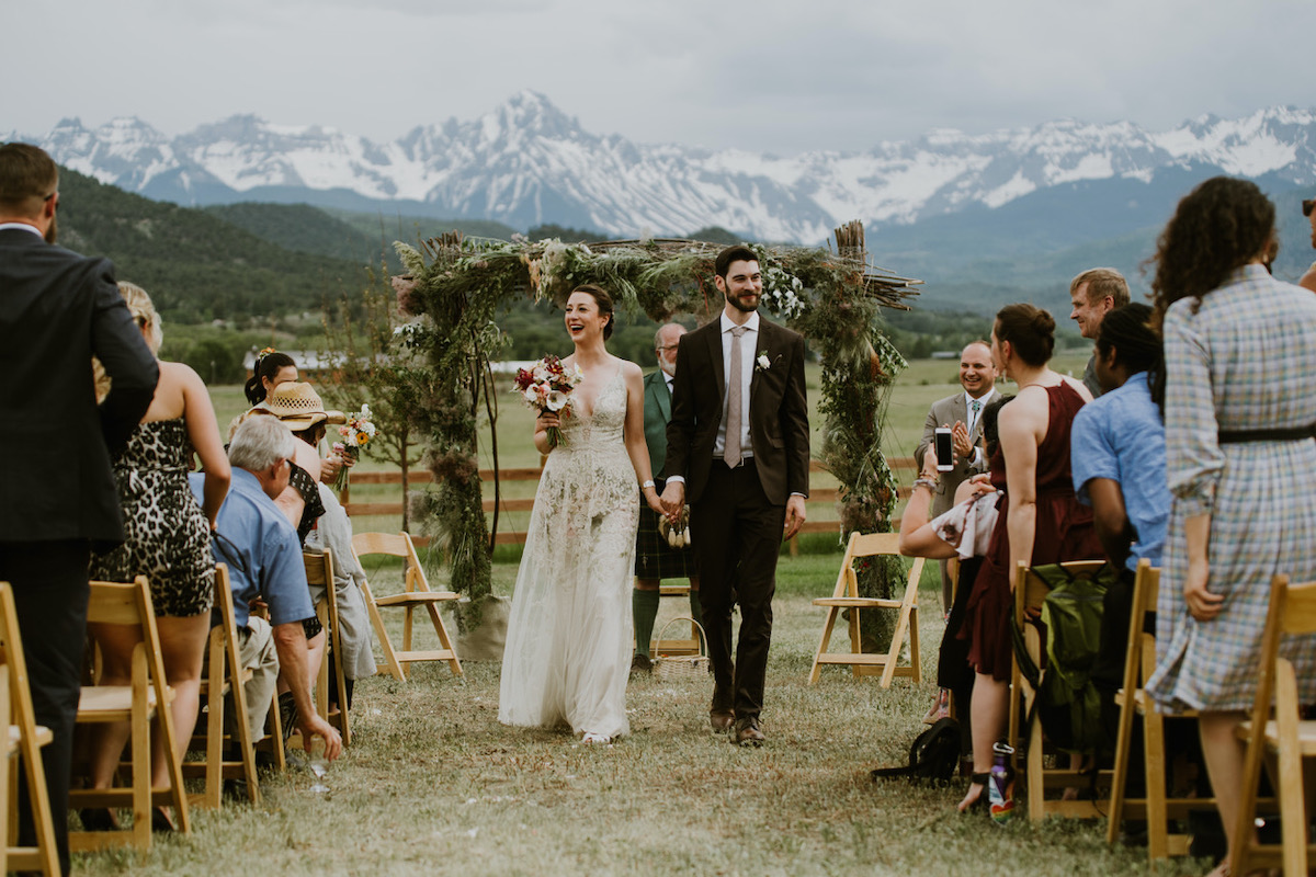 couple just wed at outdoor wedding with snowy mountain backdrop, walking back down the aisle hand in hand 